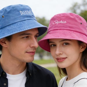 Puede incluir: Se muestran dos sombreros de pescador, uno azul y otro rosa. El sombrero azul tiene el nombre "Daniel" bordado en blanco, y el sombrero rosa tiene "Sophia" bordado en blanco. Ambos sombreros tienen un aspecto informal y desgastado.