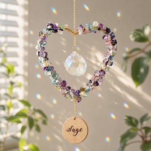 May include: A heart-shaped suncatcher featuring a wire frame adorned with small, multi-colored purple, green, and clear gemstone chips. A clear, faceted crystal teardrop hangs in the center, and a round wooden disc engraved with the word Sage hangs below.
