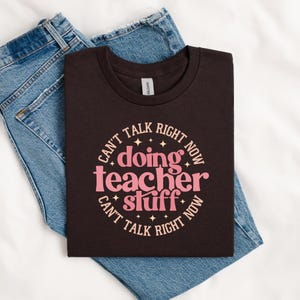 May include: A dark brown crew neck t-shirt featuring a circular graphic with the pink text "doing teacher stuff" in the centre, surrounded by the repeating beige text "CAN'T TALK RIGHT NOW" and small star accents, paired with blue denim jeans.