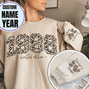Op de afbeelding: Beige sweatshirt met het jaar "1996" in een luipaardprint ontwerp. De woorden "limited edition" staan onder de cijfers. De mouw heeft de tekst "Jennifer's chapter" en een luipaardprint hart. Een cirkelvormige afbeelding bovenaan zegt "CUSTOM NAME YEAR".
