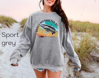 Könnte beinhalten: Ein graues Sweatshirt mit dem Aufdruck "Easily Distracted by Sharks are my favorite" auf der Vorderseite. Der Text ist in einer bunten, Retro-Schriftart gehalten.