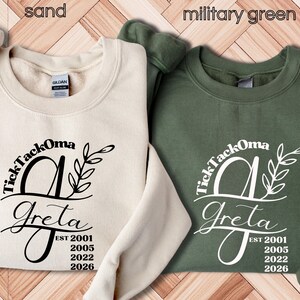 Pode incluir: Duas camisolas de gola redonda, uma cor areia e outra verde militar. Cada uma apresenta um design de monograma preto com o nome "Greta" e "TickTackOma" acima. O design inclui os anos 2001, 2005, 2022 e 2026.