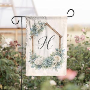 May include: A garden flag with a floral design and the letter "H" within a wooden house frame. The flag showcases a watercolour-style arrangement of blue, white, and green flowers and foliage. The flag is suspended on a black metal stand.