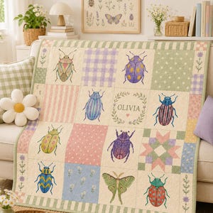 Könnte beinhalten: Eine Patchwork-Decke mit cremefarbenem Hintergrund und verschiedenen Insektenillustrationen, darunter Käfer und ein Nachtfalter, abwechselnd mit pastellfarbenen Quadraten in Vichy-Karo, Punkten und Blumenmustern. Das mittlere Quadrat zeigt den Namen OLIVIA.