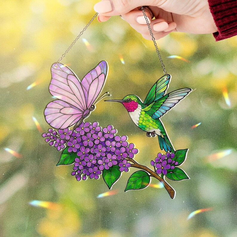 May include: A stained glass suncatcher featuring a butterfly and a hummingbird near purple flowers. The butterfly has pink wings, and the hummingbird has green and blue feathers. The suncatcher hangs from a chain.
