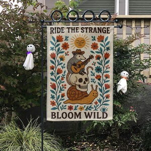 Puede incluir: Una bandera de jardín con el texto "RIDE THE STRANGE BLOOM WILD" presenta un diseño caprichoso. Representa una zarigüeya tocando la guitarra sobre una calavera y un caracol, rodeada de flores y un sol sonriente. La bandera se muestra al aire libre.