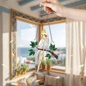 Peut inclure: Un capteur de soleil décoratif en vitrail représentant un cacatoès blanc et jaune perché sur une branche marron avec des feuilles vertes. L'oiseau est suspendu par une chaîne argentée, créant un charmant élément de fenêtre. Le capteur de soleil est tenu par une main.