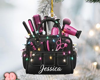 May include: A black Christmas ornament shaped like a hairstylist's bag, filled with pink hair tools. The bag is decorated with colorful lights and the name "Jessica" in white script. A gold string is attached for hanging.