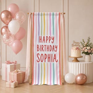 May include: A vertical rectangular banner featuring vertical stripes in pastel pink, yellow, teal, and white, with the red text "HAPPY BIRTHDAY SOPHIA" centered. The banner is displayed on a wooden stand, surrounded by pink and white balloons and gifts.