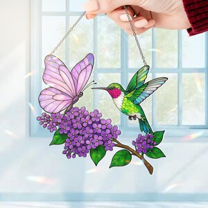 May include: Stained glass window hanging featuring a butterfly and hummingbird. The butterfly has pink wings, and the hummingbird has green and red feathers. They are positioned near a branch of purple flowers. The piece is suspended by a silver chain.