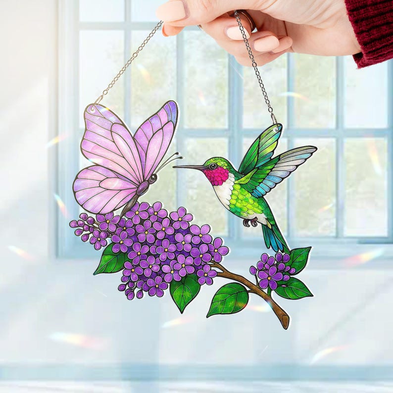May include: Stained glass window hanging featuring a butterfly and hummingbird. The butterfly has pink wings, and the hummingbird has green and red feathers. They are positioned near a branch of purple flowers. The piece is suspended by a silver chain.
