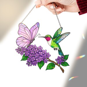 May include: Stained glass art piece featuring a butterfly and hummingbird with purple flowers. The butterfly has pink wings, and the hummingbird has green and red feathers. The art is suspended by a silver chain.