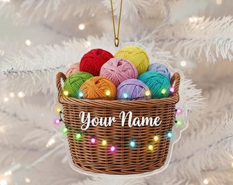 May include: A clear acrylic ornament shaped like a woven basket filled with colorful yarn balls. The ornament has the text "Your Name" and is decorated with string lights. A gold string is attached for hanging.