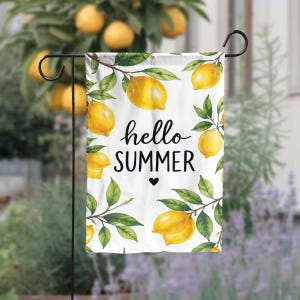 May include: A white garden flag with a watercolor design of lemons and green leaves. The flag has the words "hello SUMMER" in black script with a small black heart. The flag is hanging on a black metal stand.