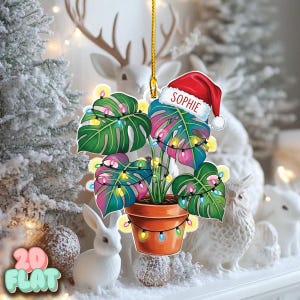 May include: A Christmas ornament featuring a potted monstera plant with colorful leaves and string lights. The ornament has a Santa hat with the name "Sophie" on it. The background includes white decorative elements.
