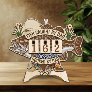 May include: Wooden fishing-themed display with the text "FISH CAUGHT BY DAD" and "HOOKED BY DAD". The display features a fish silhouette, a fishing hook, and a counter displaying "162". Decorative elements include cattails, a fishing lure, and a campfire.