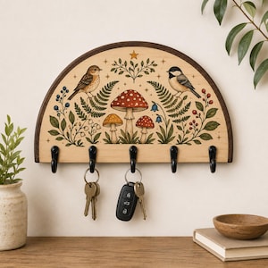 May include: A semi-circular wooden wall-mounted key holder featuring a printed illustration of two birds, red and yellow mushrooms, ferns, and berries. The light wood board has a dark wood frame and five black metal hooks, with keys hanging.