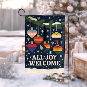May include: A navy blue garden flag with heart-shaped ornaments in various pride flag designs. The ornaments hang from a green branch with snowflakes. The text "ALL JOY WELCOME" is displayed in white.