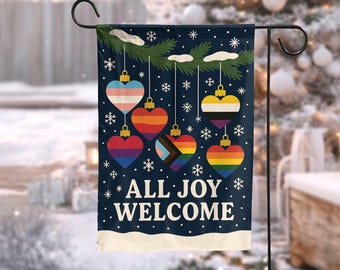 May include: A navy blue garden flag with heart-shaped ornaments in various pride flag designs. The ornaments hang from a green branch with snowflakes. The text "ALL JOY WELCOME" is displayed in white.