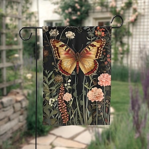 Puede incluir: Una bandera de jardín rectangular con un diseño de mariposa con alas amarillas y rojas, sobre un fondo negro con flores y follaje. La bandera se muestra en un soporte de metal negro, ideal para exteriores.