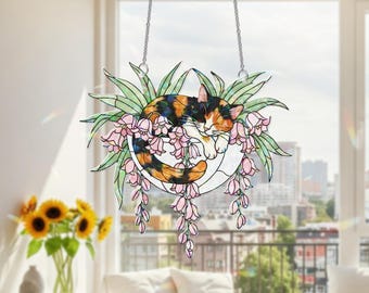 Puede incluir: Atrapasoles de gato de vidriera. Un gato calicó está acurrucado en un cuenco blanco rodeado de flores rosas y hojas verdes. El atrapasoles cuelga de una cadena plateada. El fondo muestra un horizonte urbano.