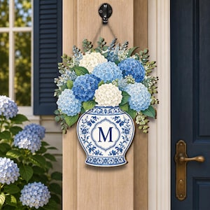 Op de afbeelding: Een decoratief deurbord met een blauw-witte vaas in porseleinstijl met de letter M gecentreerd in een kader. De vaas is gevuld met een weelderig arrangement van blauwe en witte hortensiabloesems en groene bladeren, opgehangen aan een touw.
