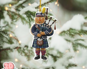 May include: A festive gingerbread man Christmas ornament playing bagpipes, dressed in a Scottish kilt and hat with the name "James". The ornament hangs from a gold string against a snowy backdrop with fairy lights.
