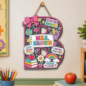 May include: A decorative wooden letter B wall hanging featuring a black background with colorful school-themed graphics, a pink bow, and a central white label reading MRS. BROWN. Additional text includes YOU'RE ENOUGH, LET'S LEARN TODAY, and YOU'RE LOVED.