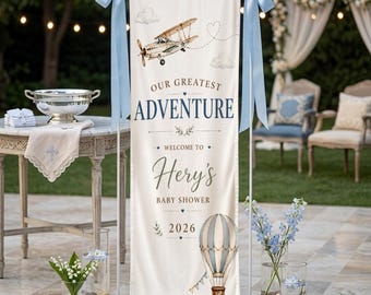 Pode incluir: Uma faixa creme com o texto "OUR GREATEST ADVENTURE" e "Welcome to Hery's BABY SHOWER 2026." Apresenta ilustrações de um biplano, nuvens e um balão de ar quente. Laços azuis claros adornam o topo.