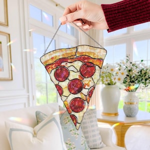 Puede incluir: Una porción de pizza de vidriera con detalles de pepperoni, corteza y queso. La porción de pizza está suspendida por una cadena. Los colores son rojo, amarillo y blanco. La porción de pizza cuelga frente a una ventana.