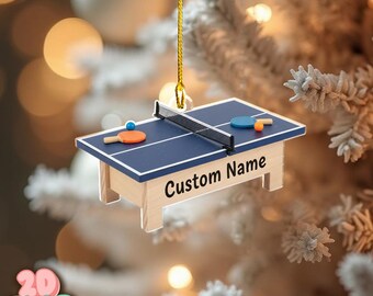 May include: A miniature ping pong table ornament with a blue surface, a black net, and two paddles and balls. The ornament has the text "Custom Name" on the side. It is hanging from a gold string.