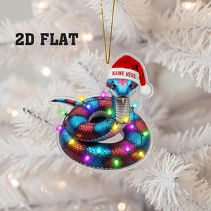 May include: A 2D flat Christmas ornament depicting a snake wearing a Santa hat and a string of colourful lights. The snake is in shades of blue, red, and purple. The text "NAME HERE" is on the hat. The text "2D FLAT" is in the upper left corner.