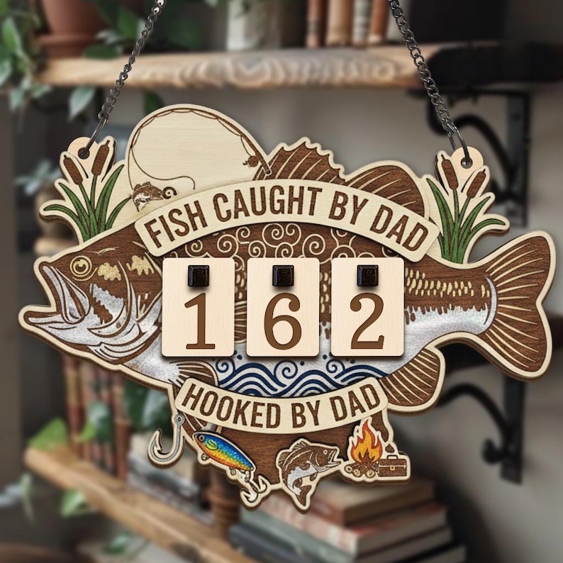 May include: Wooden fish-shaped sign with the text "FISH CAUGHT BY DAD" and "HOOKED BY DAD". The sign has a counter displaying "162" and decorative fishing-themed elements. It hangs from a chain.