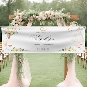 May include: A white banner with the text "Welcome to Emily's Bridal Shower" and the date 03.01.2026. The banner features illustrations of a wedding dress, rings, flowers, and a champagne glass. The banner is displayed outdoors.