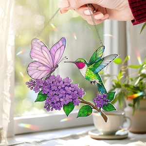 May include: A decorative window hanging featuring a butterfly and hummingbird. The butterfly has pink wings, and the hummingbird has green and blue feathers. They are positioned near purple flowers on a brown branch, suspended by a chain.