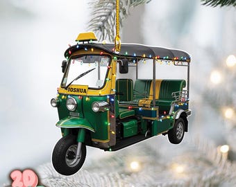 May include: A Christmas ornament shaped like a green, yellow, and black tuk-tuk, decorated with colorful string lights. The name "JOSHUA" is visible on the front. The ornament hangs from a gold string.