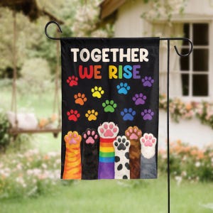 May include: Black garden flag with the words "TOGETHER WE RISE" in white and rainbow colors. The flag features colorful paw prints and illustrations of cat paws. The flag is displayed on a black metal stand in a garden setting.