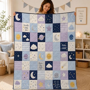 May include: A patchwork quilt featuring a celestial theme with squares in shades of blue, purple, and white. The quilt displays patterns of moons, suns, stars, planets, and clouds, with text including "twinkle little star," "moon child," and "dream big little one."