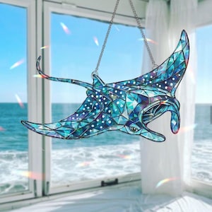 May include: A stained glass suncatcher in the shape of a manta ray, with a mosaic design in shades of blue, teal, and purple. The ray has white spots and is suspended by a silver chain. The background shows a window with a view of the ocean.