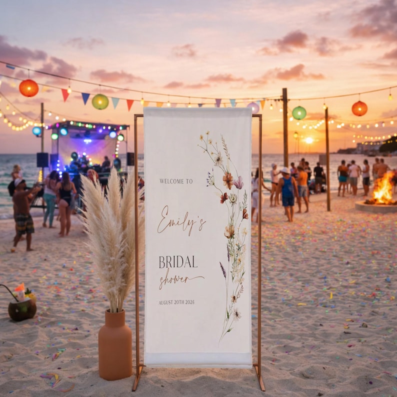 May include: A beach bridal shower banner with the text "Welcome to Emily's Bridal Shower August 20th 2026". The banner is decorated with floral illustrations and is set on a copper-colored stand. A vase of pampas grass is in front of the banner.