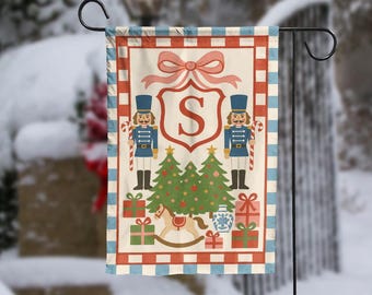 May include: A Christmas garden flag featuring a festive design. The flag displays a red and white checked border, a central shield with the letter "S", and illustrations of nutcrackers, Christmas trees, and wrapped gifts. A pink bow adorns the top.