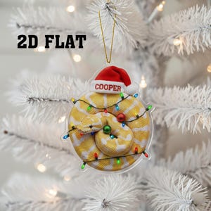May include: A clear, 2D flat Christmas ornament featuring a yellow and white snake wearing a red Santa hat with the name "COOPER". The snake is coiled and surrounded by a string of colorful Christmas lights. The text "2D FLAT" is at the top.