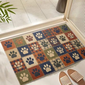 May include: A rectangular doormat with a patchwork design featuring paw prints. The mat has squares in shades of blue, orange, green, and beige, with a plaid pattern in some squares. The paw prints are in contrasting colors.