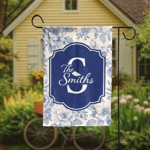 May include: A personalized garden flag featuring a blue floral pattern on a white background. The flag has a navy blue central design with the family name "The Smiths" and a large white initial "S". The flag is displayed on a black metal stand.