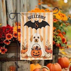 Puede incluir: Una bandera de jardín vertical con rayas beige y blancas, un gráfico de murciélago negro con el texto WELCOME y la letra S. Un cachorro golden retriever vestido de fantasma sostiene un cubo de calabaza lleno de dulces.
