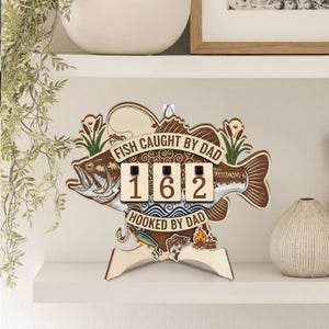 May include: Wooden fish-shaped sign with the text "FISH CAUGHT BY DAD" and "HOOKED BY DAD". The sign has three number blocks displaying "162". The sign is brown and beige with a fish illustration and fishing-themed details.