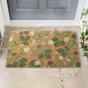 May include: Rectangular doormat with a tan background featuring a pattern of green kiwi slices, green leaves, white flowers, and brown seeds. The mat is placed in front of a gray door.