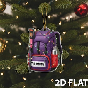May include: A purple backpack Christmas ornament with a red accent, a water bottle, and hiking poles. The ornament is decorated with a string of colorful lights and has the text "YOUR NAME" on the front. The words "2D FLAT" are at the bottom.