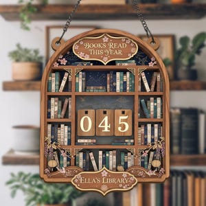 Könnte beinhalten: Holzschild in Bücherregalform mit den Texten "BOOKS READ THIS YEAR" und "Ella's Library". Das Schild hat eine Kette zum Aufhängen und zeigt ein Bücherregal-Design mit Miniaturbüchern und der Zahl "045".