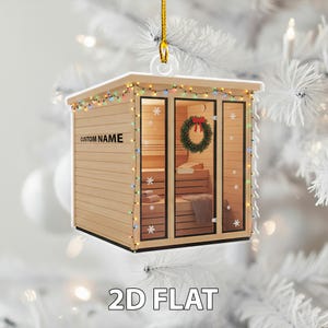 May include: A wooden sauna Christmas tree ornament, decorated with a wreath and fairy lights. The ornament has the text "CUSTOM NAME" and "2D FLAT". The sauna features glass doors and windows, and a gold hanging string.